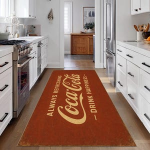 Könnte beinhalten: Ein rechteckiger, roter Coca-Cola-Küchenteppich mit dem Coca-Cola-Logo in cremefarbenem Text. Der Teppich trägt die Aufschriften "ALWAYS REFRESHING" und "DRINK HAPPINESS". Der Teppich liegt auf einem Holzboden in einer Küche.