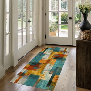 May include: A rectangular area rug with an abstract geometric design in shades of teal, orange, yellow, gray, and black. The rug is placed on a wooden floor in a hallway, with white doors and natural light visible in the background.
