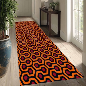 May include: A long, rectangular rug with a repeating geometric pattern of orange, black, and burgundy hexagons. The rug is placed in a hallway with wooden floors, a large potted plant, and a dark wooden table.