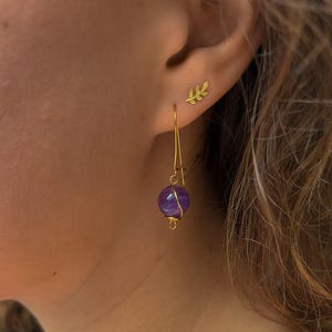 Op de afbeelding: Een close-up van een oor met twee oorbellen. Eén is een kleine gouden bladknop. De andere is een lange, gouden draad oorbellen met een ronde, paarse amethist kraal. De oorbellen zijn geplaatst tegen de huid en het haar van de persoon.