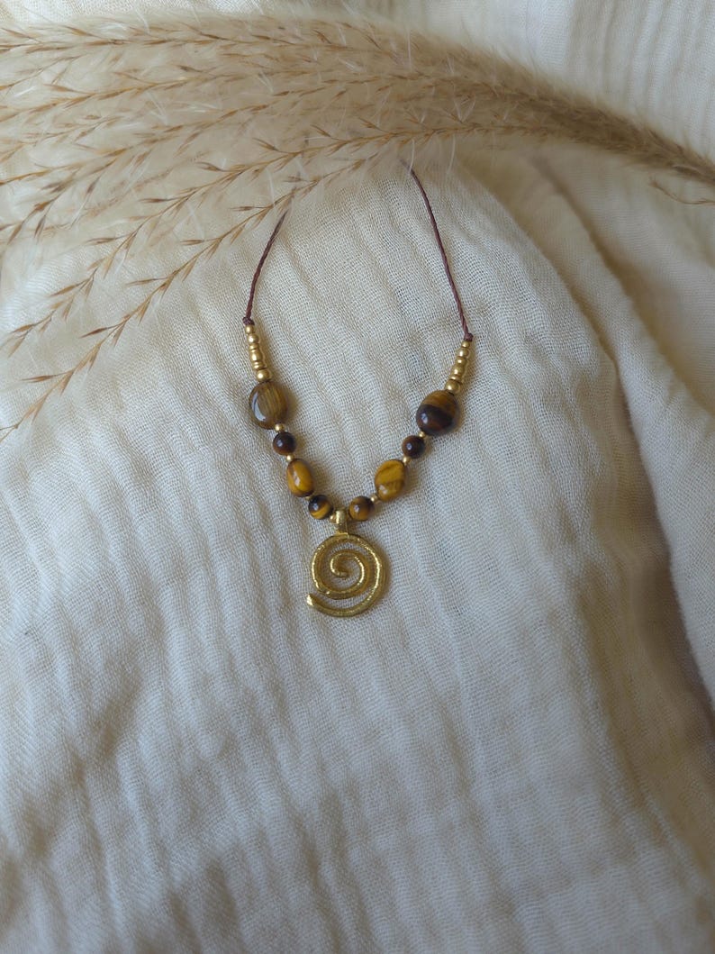 May include: A necklace featuring a gold-toned spiral pendant suspended from a thin brown cord. The design includes a series of polished tiger eye beads in shades of brown and gold, interspersed with small metallic gold-toned spacer beads.