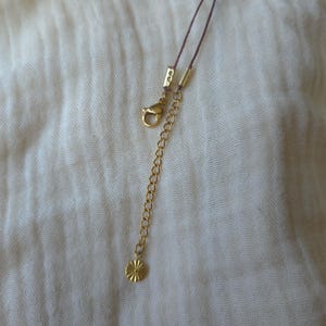 May include: A thin brown cord necklace featuring a gold-toned metal lobster claw clasp, a gold-toned metal extender chain, and a small, round, gold-toned metal charm with a sunburst or star-like textured pattern at the end of the chain.