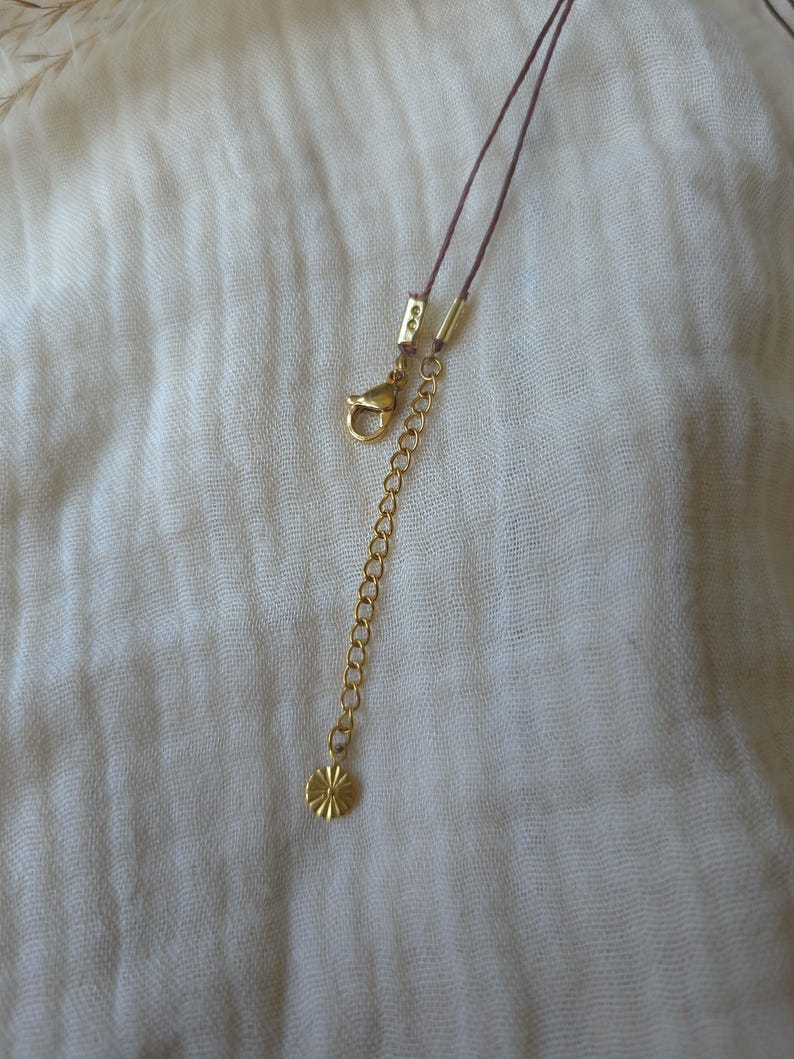 May include: A thin brown cord necklace featuring a gold-toned metal lobster claw clasp, a gold-toned metal extender chain, and a small, round, gold-toned metal charm with a sunburst or star-like textured pattern at the end of the chain.