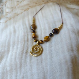 May include: A necklace featuring a gold-toned spiral pendant suspended from a thin cord. The design includes a series of brown and gold tiger eye beads interspersed with small metallic gold-colored spacers arranged symmetrically along the cord.
