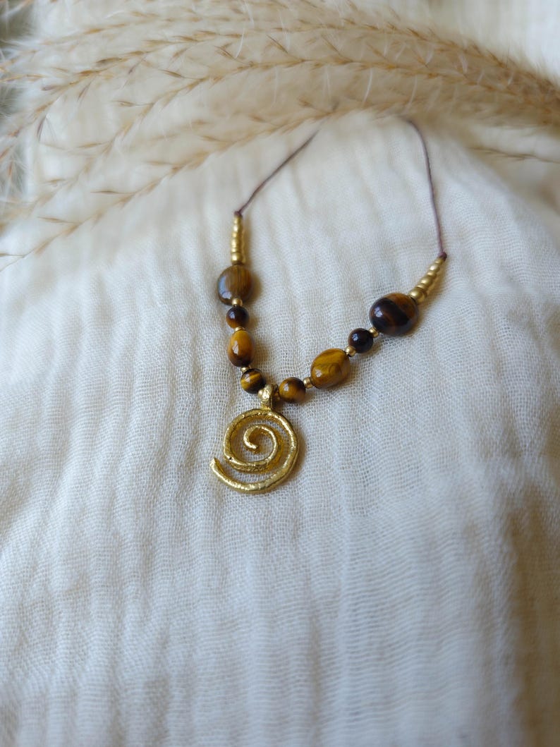 May include: A necklace featuring a gold-toned spiral pendant suspended from a thin cord. The design includes a series of brown and gold tiger eye beads interspersed with small metallic gold-colored spacers arranged symmetrically along the cord.