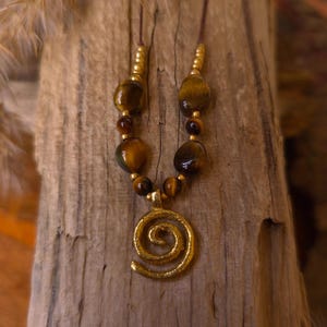 May include: A necklace featuring a gold-toned spiral pendant suspended from a brown cord. The design includes polished, amber-colored tiger eye beads interspersed with small, metallic gold-toned spacer beads arranged symmetrically along the cord.