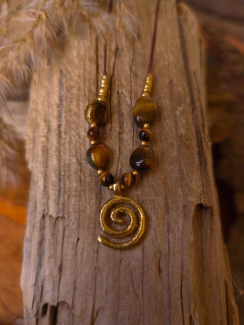 May include: A necklace featuring a gold-toned spiral pendant suspended from a brown cord. The design includes polished, amber-colored tiger eye beads interspersed with small, metallic gold-toned spacer beads arranged symmetrically along the cord.