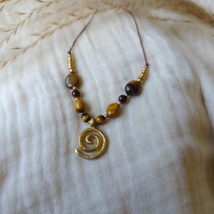 May include: A necklace featuring a gold-toned spiral pendant suspended from a thin cord. The design includes a series of brown and gold tiger eye beads interspersed with small metallic gold-colored spacer beads arranged symmetrically along the cord.