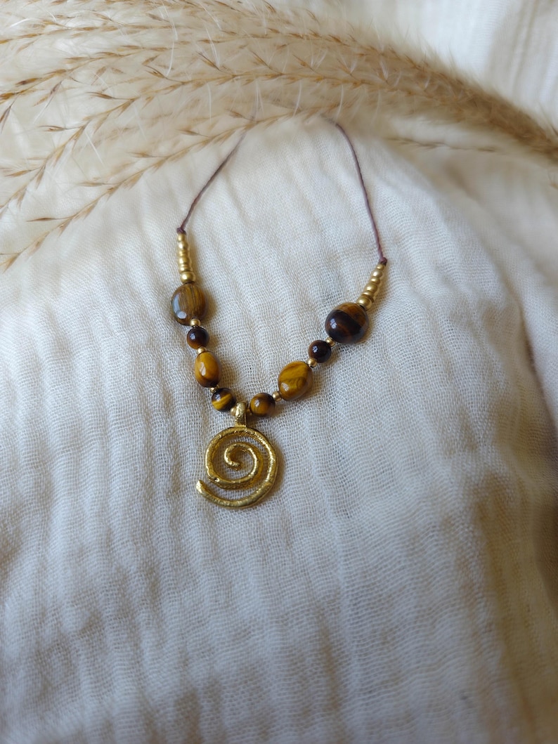 May include: A necklace featuring a gold-toned spiral pendant suspended from a thin cord. The design includes a series of brown and gold tiger eye beads interspersed with small metallic gold-colored spacer beads arranged symmetrically along the cord.