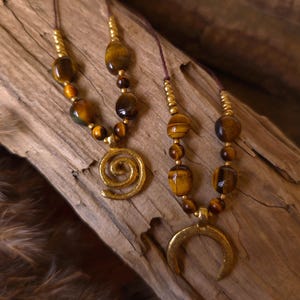May include: Two necklaces featuring brown and gold tiger eye beads and small metallic gold beads on thin brown cords. One necklace has a gold-toned spiral pendant, while the other features a gold-toned crescent moon-shaped pendant.