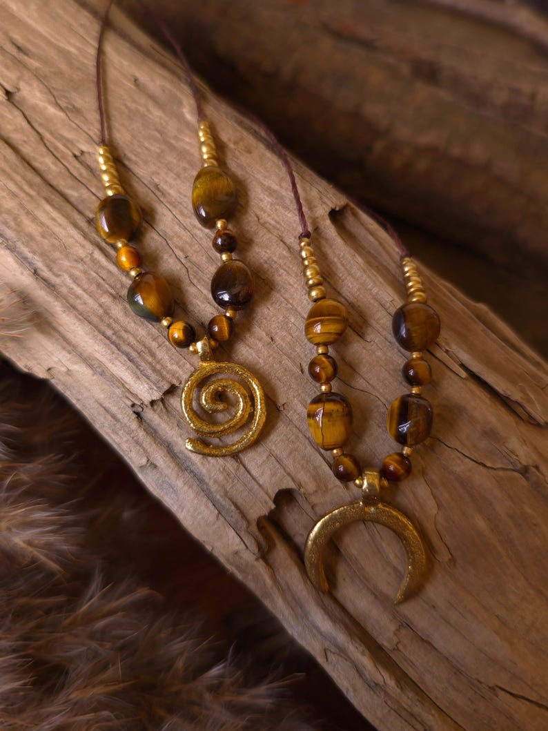 May include: Two necklaces featuring brown and gold tiger eye beads and small metallic gold beads on thin brown cords. One necklace has a gold-toned spiral pendant, while the other features a gold-toned crescent moon-shaped pendant.