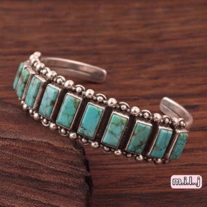 May include: A silver cuff bracelet featuring a row of rectangular turquoise stones with dark matrix patterns. The bracelet is bordered by a beaded silver texture along the top and bottom edges, creating a detailed and structured metal design.