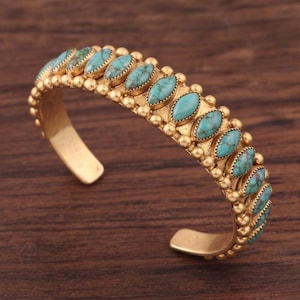 May include: A gold-toned cuff bracelet featuring a row of marquise-shaped turquoise stones with dark veining. The metal band is decorated with small, round gold beads bordering the stones, creating a textured, open-ended circular design.