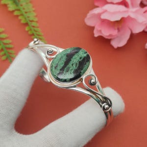 May include: A silver bracelet with an oval green and black stone. The bracelet has a decorative, open design with swirling details. The stone is set in a silver bezel. The bracelet is displayed against a soft background.