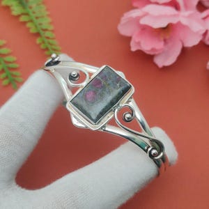 May include: A silver-toned cuff bracelet featuring a rectangular, dark gray stone with red inclusions. The bracelet has an open design with decorative swirls and small ball accents, showcasing a blend of modern and classic styles.