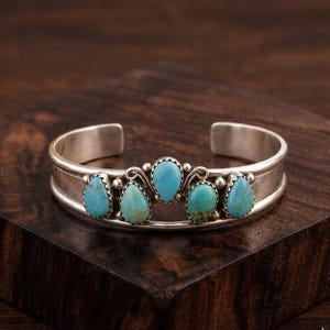 May include: A silver cuff bracelet featuring five teardrop-shaped turquoise stones arranged in a row. The stones are set in smooth silver bezels with small silver bead accents between them. The band is a solid, polished silver open-ended cuff design.