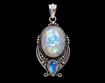 Puede incluir: Un colgante de plata con una gran piedra de luna ovalada con destellos iridiscentes azules y amarillos, situada sobre una piedra de luna más pequeña en forma de pera. Las piedras están engastadas en un marco de plata con detalles de alambre trenzado.