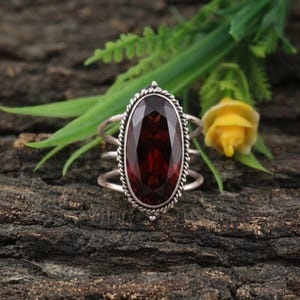 Pode incluir: Um anel de prata com uma grande pedra preciosa granada de corte oval e vermelho profundo, engastada numa borda de metal com design de corda torcida. O aro consiste em três fios de prata finos, lisos e separados que convergem nas laterais.