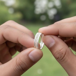 May include: A silver ring with an openwork pattern. The ring is held between two hands against a blurred green backdrop. The ring features a repeating design of small circles around the band.