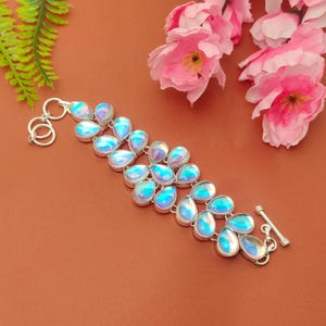 Puede incluir: Una pulsera de plata con piedras iridiscentes en forma de lágrima. Las piedras son azules y moradas, engastadas en plata. La pulsera tiene un cierre de palanca. Se muestra sobre una superficie marrón.