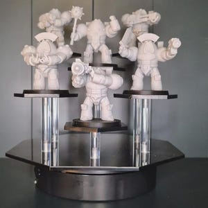 Puede incluir: Una exhibición con seis figuras de robots en miniatura blancas, cada una con diferentes armas y accesorios. Las figuras están montadas sobre plataformas negras sostenidas por columnas de acrílico transparente, dispuestas sobre una base giratoria negra.