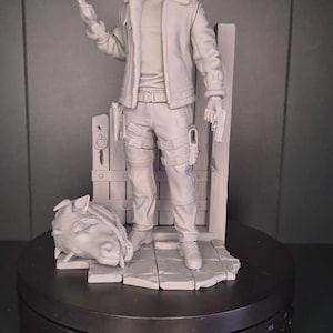 Può includere: Statuetta grigia dettagliata di un personaggio che tiene in mano un'arma e una pistola. La figura indossa una giacca, pantaloni e stivali. Una testa di cavallo è alla base. La statuetta è su una piattaforma rotante.