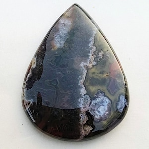 Könnte beinhalten: Ein polierter, tropfenförmiger Edelstein mit dunkler, glänzender Oberfläche. Der Stein weist eine Mischung aus Farben auf, darunter tiefes Schwarz, Lila und Hauch von Weiß und Braun, wodurch ein einzigartiges, marmoriertes Muster entsteht. Der Edelstein wird wahrscheinlich für Schmuck oder Dekorationszwecke verwendet.