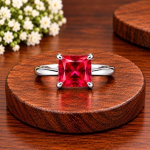 Puede incluir: Un anillo de tono plateado con una piedra preciosa roja vibrante de corte cuadrado, sujeta por cuatro garras. La banda es lisa y pulida, mostrando un diseño limpio que resalta el color carmesí intenso de la piedra central.