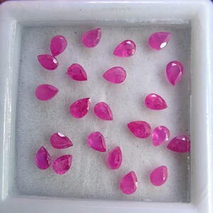 Könnte beinhalten: Eine Sammlung facettierter Rubine in Tropfenform. Die Edelsteine haben eine tiefrosa-rote Farbe und werden in einer weißen, rechteckigen Schale mit weichem, weißem Innenraum präsentiert. Die Edelsteine variieren leicht in Größe und Form.