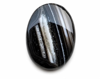 Könnte beinhalten: Ein polierter, oval geformter Stein mit glänzender Oberfläche. Der Stein weist ein auffälliges Muster aus schwarzen, weißen und grauen Bändern und Wirbeln auf. Der Edelstein ist vor einem schlichten weißen Hintergrund platziert, was seine natürliche Schönheit hervorhebt.