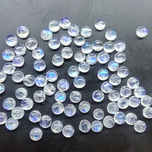 Könnte beinhalten: Eine Ansammlung kleiner, runder, klarer Mondstein-Cabochons. Jeder Stein hat ein durchscheinendes Aussehen mit einem subtilen Spiel von blauem und weißem Licht, das über eine dunkle Oberfläche verstreut ist. Diese polierten Edelsteine sind ideal für die Schmuckherstellung.
