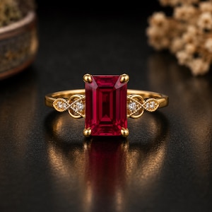 Könnte beinhalten: Ein Goldring mit einem großen, rechteckigen, tiefroten Edelstein im Smaragdschliff als Mittelstein. Die Ringschiene zeigt auf beiden Seiten filigrane Metallarbeiten, die mit kleinen, runden, klaren weißen Steinen besetzt sind.