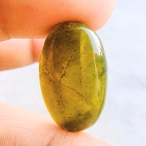 Könnte beinhalten: Ein oval geschliffener, polierter Edelstein mit transluzentem, olivgrünem Farbton. Der Stein hat eine glatte, glänzende Oberfläche und subtile innere Muster. Der Edelstein wird zwischen zwei Fingern gehalten und zeigt seine natürliche Schönheit und Klarheit.