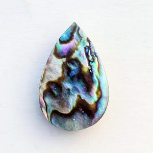 Könnte beinhalten: Ein tropfenförmiger Cabochon aus Abalone-Muschel mit schillernden Farben wie Blau, Lila, Grün und Braun. Die polierte Oberfläche zeigt ein wirbelndes Muster, das die natürliche Schönheit der Muschel hervorhebt. Geeignet für die Schmuckherstellung.