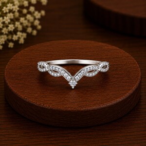 Könnte beinhalten: Ein silberfarbener V-förmiger Ring mit einem zarten, gedrehten Banddesign. Die gesamte Oberfläche des Rings ist mit kleinen, klaren, funkelnden Steinen besetzt, wobei ein einzelner, etwas größerer runder Stein in der Mitte des V-förmigen Punktes sitzt.