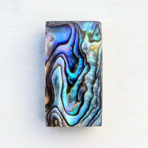 Könnte beinhalten: Rechteckige Abalone-Schale mit schillernden blauen, grünen und violetten Wirbeln. Die polierte Oberfläche reflektiert das Licht und zeigt die natürlichen Muster der Schale. Die Form der Schale ist ein Rechteck.