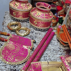 May include: A collection of decorative items in pink and gold. The image shows two ornate, lidded vessels, gold rings, decorative fans, and long cylindrical objects. The items have detailed gold patterns and are arranged on a textured surface.