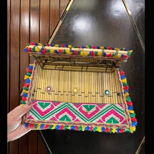 Peut inclure: Une boîte rectangulaire en bambou avec un couvercle à charnière, ornée de pompons colorés et de motifs géométriques brodés en rose, vert et blanc. La boîte comporte de petits accents miroir et est probablement utilisée pour le rangement.