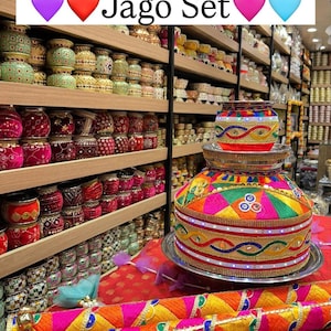 May include: A vibrant Jago Set featuring detailed embroidery and decorative elements. The set comprises a large, ornamented pot and two decorative rolls, presented on a red surface. Shelves filled with various decorative items are also visible.