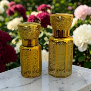 May include: Two gold-colored glass perfume bottles with hexagonal bodies and intricate embossed patterns. Each bottle features a matching metallic gold cap with a detailed filigree design, standing upright with a reflective finish.