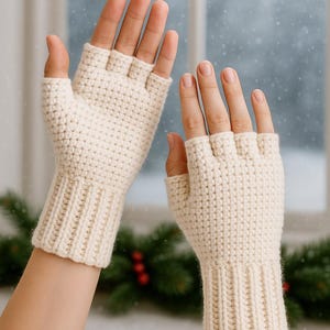 Può includere: Guanti senza dita color crema, lavorati a mano all'uncinetto con polsino a coste. I guanti sono progettati per coprire la mano fino alle nocche, lasciando libere le dita. Lo sfondo mostra una finestra innevata e rami sempreverdi.