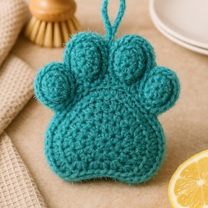 May include: A teal crocheted paw-shaped dish scrubby with a loop for hanging. The scrubby is made of textured yarn and is surrounded by a wooden brush, a folded towel, a lemon slice, and a stack of white plates.