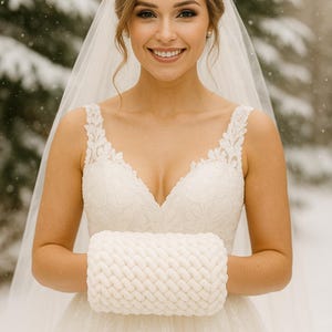 Pode incluir: Uma noiva em um vestido de noiva de renda branca e véu sorri para a câmera. Ela segura uma luva branca de malha. O fundo é uma floresta nevada, criando um tema de casamento de inverno.