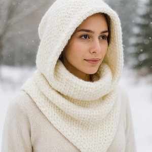 Puede incluir: Conjunto de capucha y bufanda de punto color crema. La capucha enmarca la cara y la bufanda cae alrededor del cuello. El tejido de punto grueso proporciona calidez. El fondo es una escena nevada al aire libre.