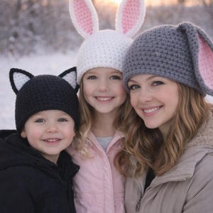 May include: Three people wearing handmade animal ear hats. A child wears a black cat ear hat, another wears a white bunny hat with pink inner ears, and an adult wears a gray bunny hat with pink inner ears. All are smiling in a snowy outdoor setting.