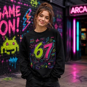 May include: A black long-sleeve sweatshirt featuring a vibrant graphic print of the number 67 in neon green and pink. The design includes retro arcade elements like pixelated game consoles, joysticks, stars, and lightning bolts with text.