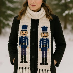 Puede incluir: Bufanda color crema con flecos con dos figuras de cascanueces bordadas con uniformes azules. La bufanda se lleva sobre un abrigo negro y se combina con un gorro verde oscuro. El fondo es una escena nevada al aire libre.