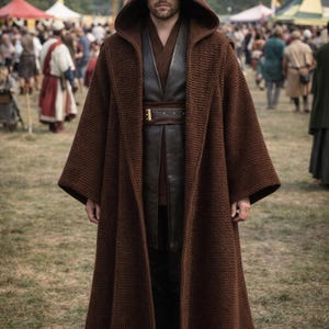 May include: A brown hooded cloak with wide sleeves, worn over a dark leather vest and belt. The cloak appears to be made of a knitted material. The person is standing in a grassy field with a crowd in the background.