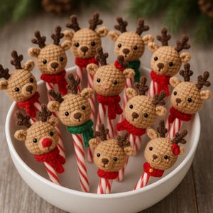 Puede incluir: Un cuenco blanco lleno de bastones de caramelo rematados con cabezas de reno de ganchillo. Los renos tienen cornamentas marrones, ojos negros y llevan bufandas rojas o verdes. Un reno tiene la nariz roja y otro un lazo rojo.