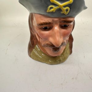 May include: A ceramic mug shaped like a man's head, featuring a gray hat with gold crossed sabers. The mug has a detailed face with a mustache and long brown hair, and a green collar.